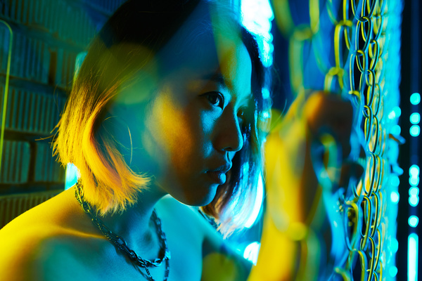 A young woman in urban style with short dyed hair and a necklace stands behind a metal mesh fence holding on to it with her hands and lit by yellow and blue light