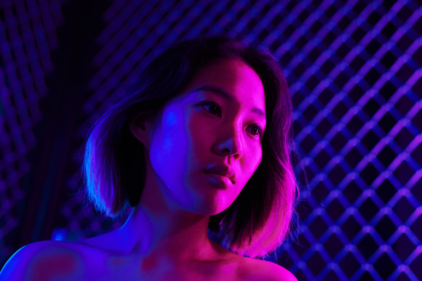 A female with short hair and dyed tips stands against the background of a metal grid in neon blue and pink light turning her head to the side