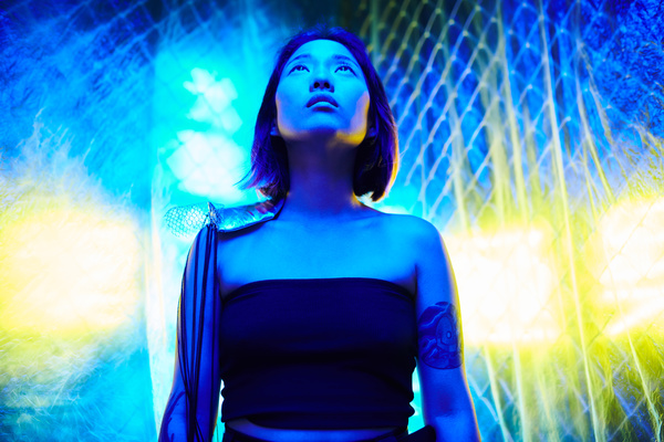 A woman with a short haircut in a black crop top without straps and with a tattoo on her shoulder stands in the rays of cold tones and looks up against the background of a metal mesh and yellow glare of light