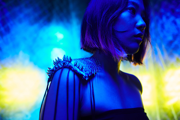 A girl with a square haircut and dyed hair tips and with a shoulder pauldron stands in a half-turn illuminated by blue light against the background of a chain-link fence and yellow lights