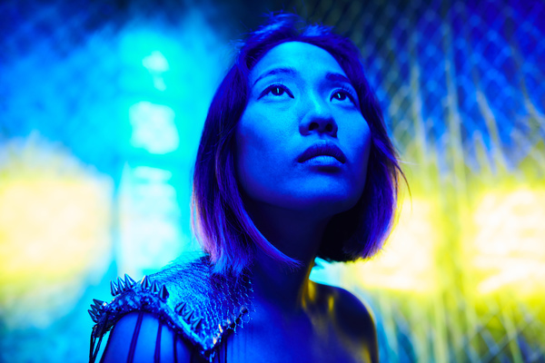 Illuminated by blue neon light female with a square haircut and her head held up looks up wearing a shoulder pauldron is against a background of metal mesh and yellow light