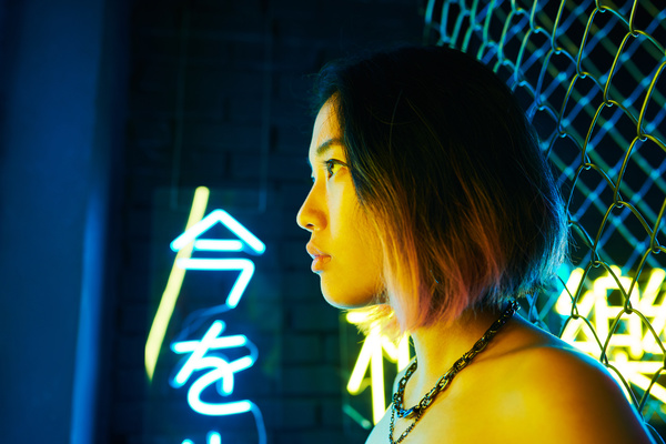 The profile of a short-haired girl illuminated by yellow light with a necklace around her neck standing with her back to a metal grid with hieroglyphs illuminated by blue light on the background