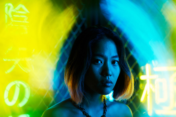A girl with a bob haircut and a chain around her neck stands under cold light with flashes of blue and yellow neon near her face against a backdrop of a chain-link fence and illuminated symbols