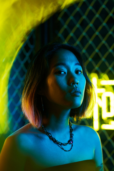 A girl with short dyed hair and an accessory around her neck with her head slightly raised standing in the cold light with flashes of yellow neon in her face against a backdrop of fence grid and illuminated hieroglyphics