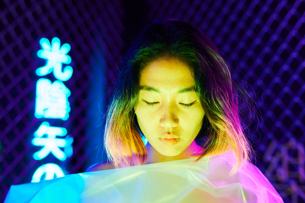 A woman with short dark hair dyed at the tips covered herself with cellophane material with her head down and eyes closed under neon blue and pink light against a background of mesh and blue hieroglyphs