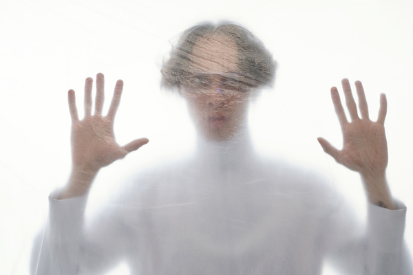 A blurred image of a man with a voluminous hairstyle dressed in white through a cellophane covering looking straight ahead with his palms raised