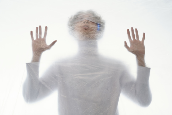 A blurry image of a curly-haired man in white clothes through a cellophane coating looking up with his palms raised standing on a white background