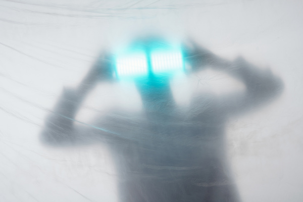 A blurred image through the cellophane covering of a male dressed in white holding sources of bright blue light with both hands at his face