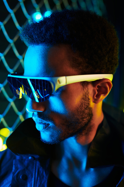Portrait of a black-haired man with a beard wearing black cyberpunk-style sunglasses with white frames dressed in a black jacket looking away lit by blue neon light against a metal grid
