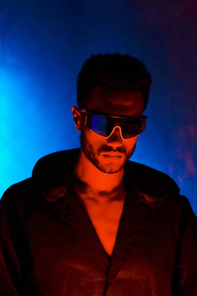 A male cyberpunk with short black hair and stubble wearing stylish glasses wearing a black hooded jacket looks down with a red-lit face standing on a neon blue background