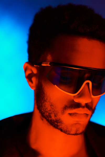 A close-up of a cyberpunk man with short dark hair and bristles in fashionable glasses looking down with his face illuminated by red rays on a neon-blue background