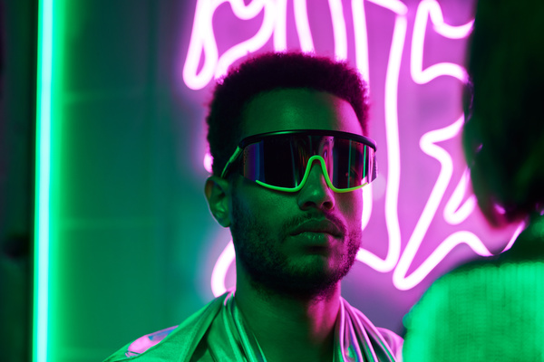 Close-up of a cyberpunk-inspired man with short dark hair and stubble wearing fancy glasses looking at the girl opposite with neon pink signs in the background and green light