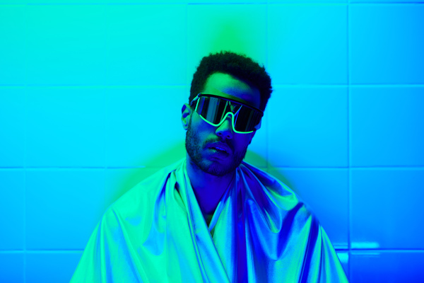 A cyberpunk man with short curly black hair and a beard wearing stylish glasses and a glossy robe stands against a tiled wall in neon light of cyan