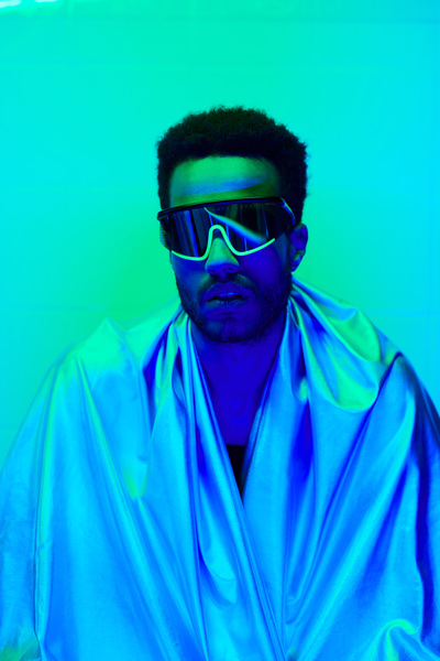 A male short haired cyberpunk with and a beard wearing fashionable single-lens sunglasses and a shining cloak standing against a wall in a room with neon blue-green lighting