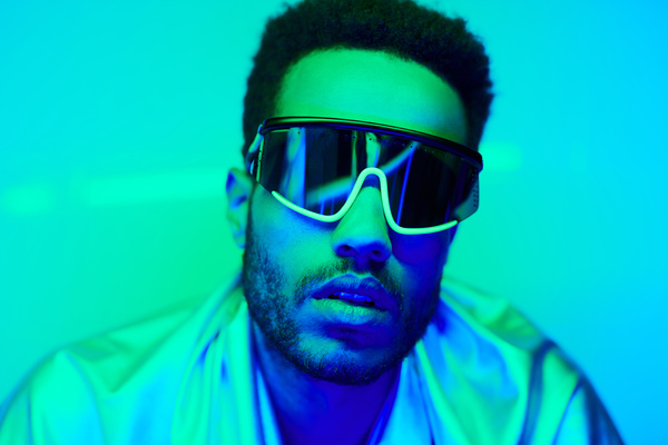 Close-up of a dark-haired young man with bristles wearing fancy black-rimmed sunglasses and a garment of glossy fabric illuminated by neon turquoise light