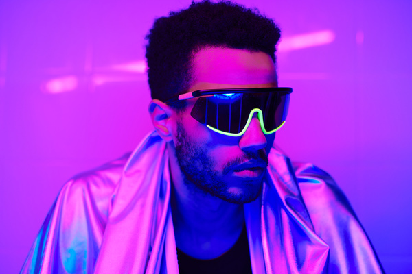 A male cyberpunk peson with black curly hair and a short beard wearing a reflective cape over a black T-shirt and in dark-rimmed glasses looks away in a room with pink neon lighting
