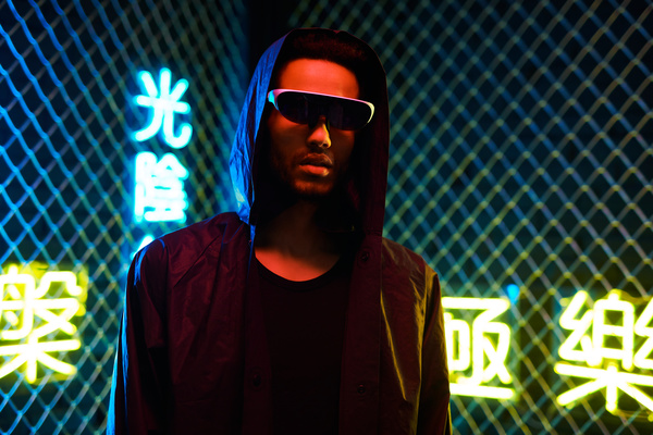 A young cyberpunk man with dark hair and a beard wearing a single-lens black sunglasses with white frames and a black hooded jacket over a black T-shirt stands against a metal mesh fence and yellow and blue neon symbols