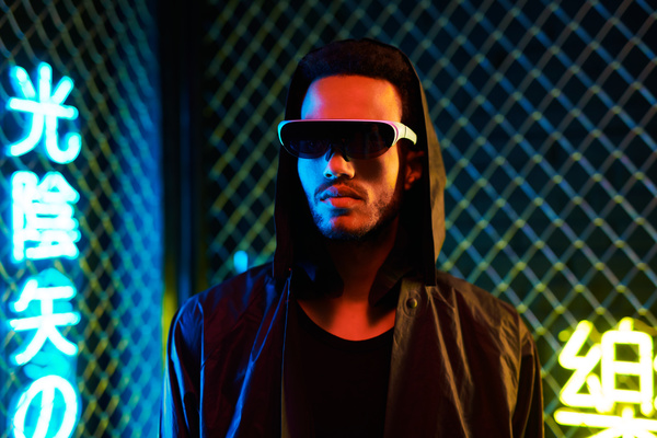 Close-up of a young cyberpunk man with black hair and a beard wearing a single-lens black sunglasses with white frames dressed in dark-colored clothes is against a metal mesh fence and yellow and blue neon hieroglyphics