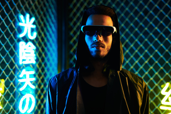Close-up of a cyberpunk black haired man with beard in a black sunglasses with one lens and white frame dressed in black attire with a hood is against a metal mesh and yellow and blue lilluminated hieroglyphics