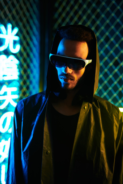 A young man with black hair and a beard wearing single-lensed black cyberpunk-style sunglasses dressed in dark-colored clothing stands half-turned against a metal-mesh fence and yellow and blue neon hieroglyphics