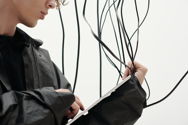 A man in cyberpunk style dressed in a black jacket standing sideways and holding a white tablet grabbing a couple of wires that dangle around him on a white background