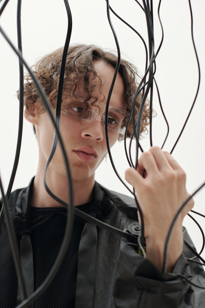 A cyberpunk man with auburn curly hair and a cross-shaped earring wearing glasses with a cyber pattern dressed in black with a serious expression on his face grabs some black wires that come from the sleeves of his jacket