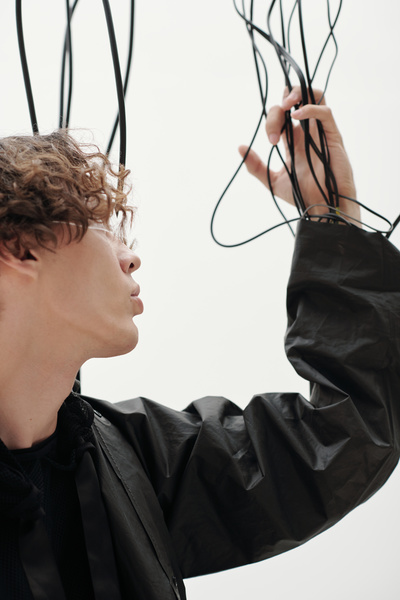 A cyberpunk man with brown wavy hair wearing clear cyber-patterned single-lens glasses dressed in a black outfit standing against a white background holding several black wires coming out of the sleeve of his jacket and looking at them