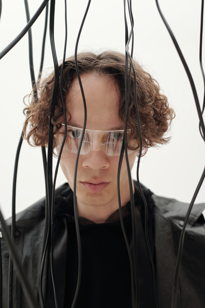A cyberpunk man with brown curly hair wearing clear glasses with a one-lens and a cyber pattern dressed in black with a strong expression standing against a light background with black cords hanging down his face and around him
