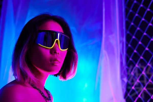 A short-haired woman in fashionable glasses in black and yellow rim and with a chain around her neck stands half-turned illuminated by pink neon with a plastic film over a mesh fence in the rays of blue light behind her