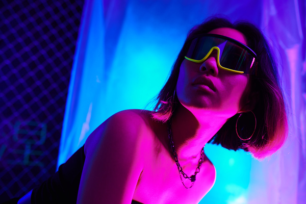 A woman with short hair in cyber glasses and an earring in her ear and a necklace around her neck leaned half-turned under pink light against a background of polymer film and a mesh fence illuminated with blue neon