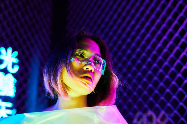 A young woman with short dyed hair at the ends wearing transparent glasses with one lens and a cyber pattern on them looks away hiding behind a light cellophane film while in a room with neon lighting metal fencing and blue illuminated hieroglyphs