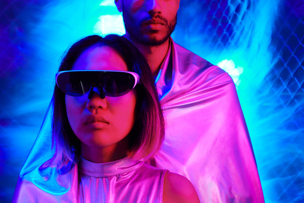 A woman in white-rimmed sunglasses dressed in a suit of shining fabric with her face illuminated with red neon light stands with a man behind her who is wearing a cloak of the same fabric against the background of a cellophane coating