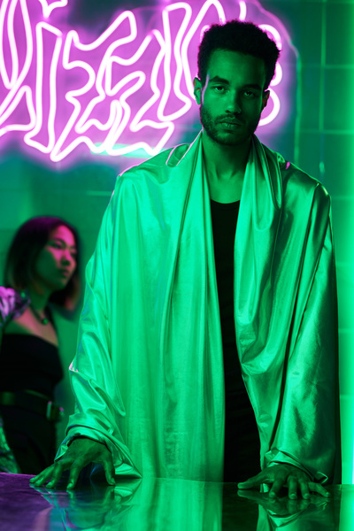 A cyberpunk guy with a short afro in a reflective cloak with his hands on a mirrored table stands in a room with green lighting and neon pink graffiti