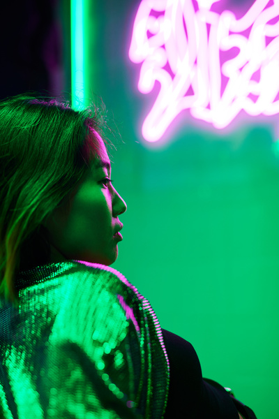 The profile of a short-haired girl in a blouse shimmering in the light sitting in a bar with green light and neon inscriptions illuminating her face with pink beams
