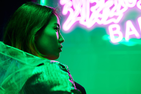 Profile of a girl with a bob haircut in a shiny jacket with a see-through hood sitting in a green-lit bar with neon pink writing on the wall behind her