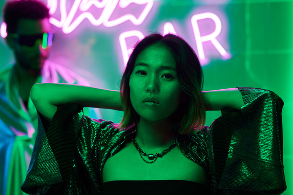 Dressed in reflective clothing with wide sleeves and a chain around her neck a woman with a short haircut stands with her hands behind her head in neon light against a background of pink symbols