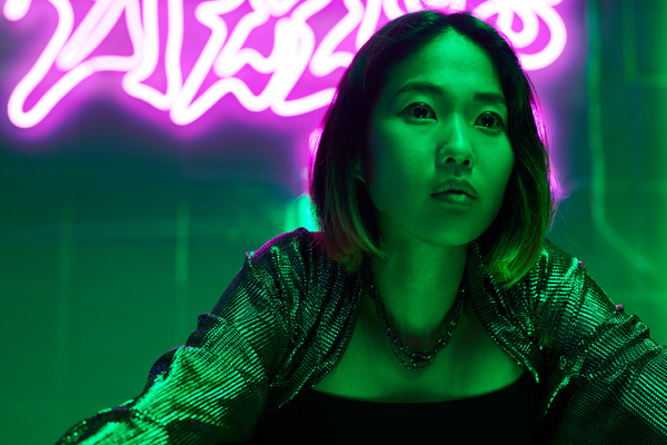 A young female with a short haircut wearing a glittering bolero over a dark top with a chains necklace around her neck looks aside leaning on a surface against a neon signs in a room lit by green rays