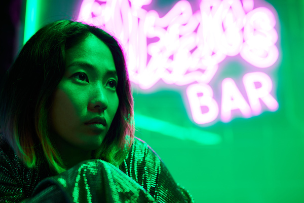 Profile of a woman with short black hair in a shiny blouse sitting in a room with green lighting and neon signs leaving pink light highlights on her face