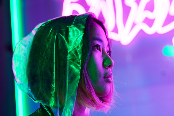 A woman with a short haircut in a transparent hood illuminated by pink light against the background of neon signs looking straight