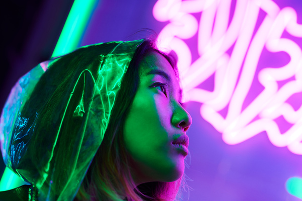 A woman with short hair in a translucent hood in the rays of green and pink neon standing in front of glowing symbols and looking up
