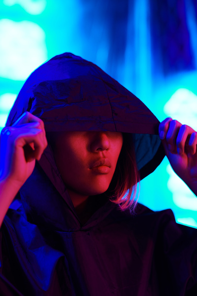 A woman with short hair lowered the hood of her black raincoat over her eyes while standing under neon red lighting on a background illuminated by blue rays