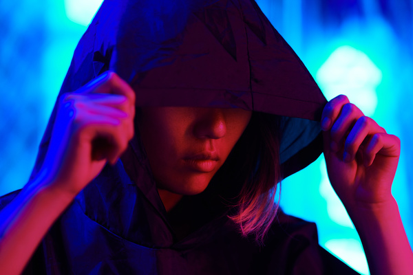 A woman with short dyed hair hides her eyes with the hood of her dark robe with her face and hands illuminated by red light