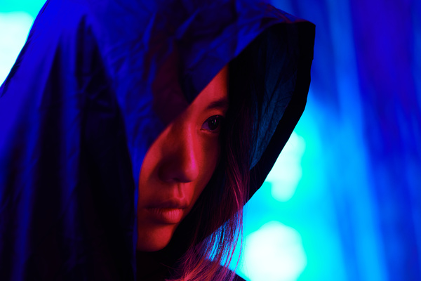 A woman looks out from under the hood of her black clothes hiding half of her face illuminated by a bright scarlet light against a background illuminated by blue rays