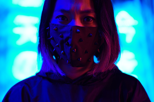 A young female with short black hair and pink tips wearing a dark colored spiked mask in a black hoodie looks straight ahead with a stern expression standing under the neon blue-red lighting