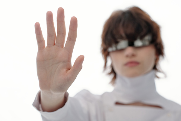 A young futuristic-looking female with short brown hair wearing silvery with one lens glasses and fashionable white outfit stands with her arm outstretched