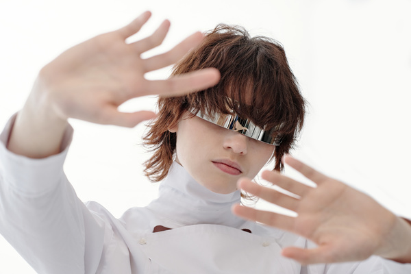 A young futuristic-looking female with short brown hair wearing silvery with one lens glasses and fashionable white outfit stands with both arms outstretched forward