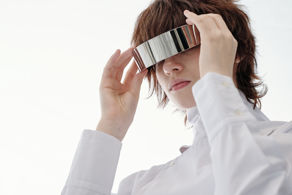 A futuristic-looking young woman with short brown curly hair wearing a modern white outfit removes her single-lens metal glasses
