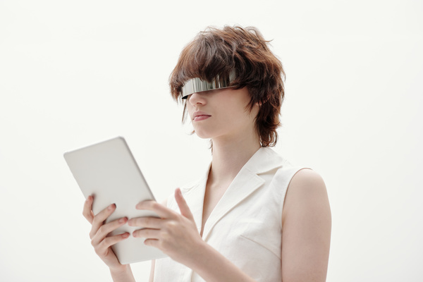 A cyberpunk woman with brown tousled hair in silvery sunglasses wearing a white sleeveless blouse with a double-breasted collar standing against white background holds a white tablet