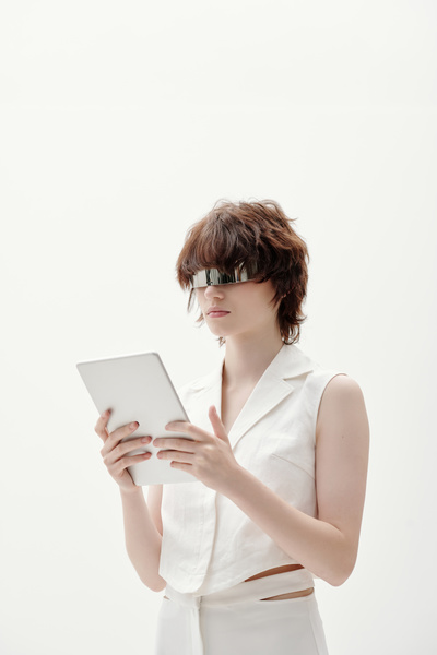 A cyberpunk female with auburn tousled hair in silvery glasses wearing a white sleeveless blouse with a double-breasted collar and pants with slits on the pockets standing sideways against white background holds a white tablet