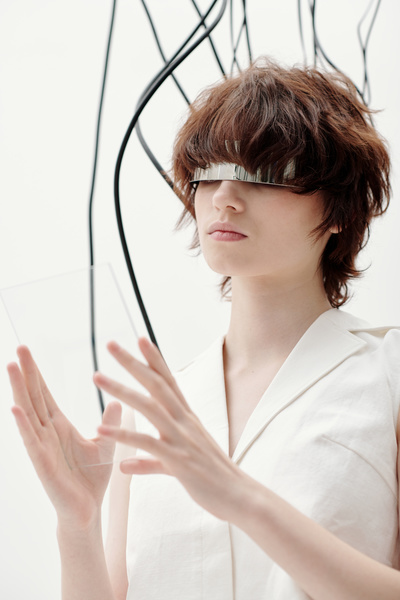 A female cyberpunk with a short haircut wearing silver single-lens glasses and a white sleeveless blouse holding a see-through rectangular screen with both hands stands among hanging dark cables on a white background